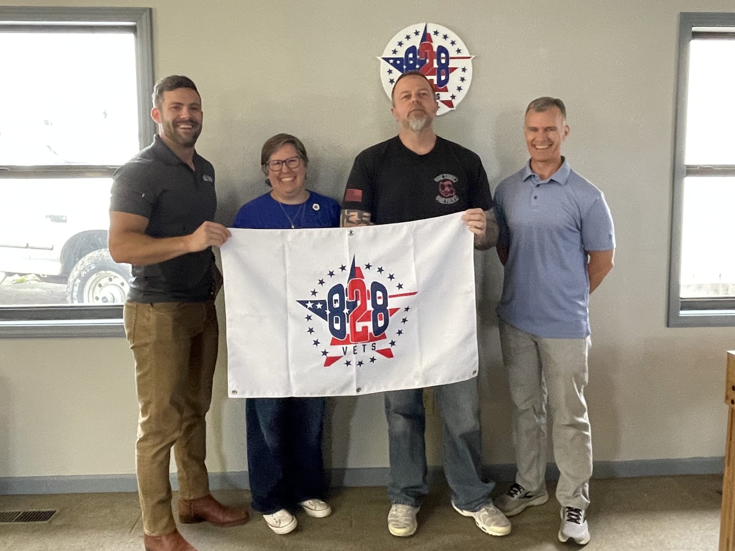 Macon County Community Foundation board member Kevin Holland stands with Tyler Cloer, Starlett Henderson, and Craig “Mac” McManus at 828 Vets headquarters, holding an 828 Vets flag