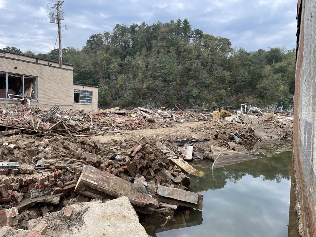 Image of downtown Marshall, NC, after Hurricane Helene. Image depicts rubble covering a street, with floodwaters still at least several feet high next to two damaged buildings.