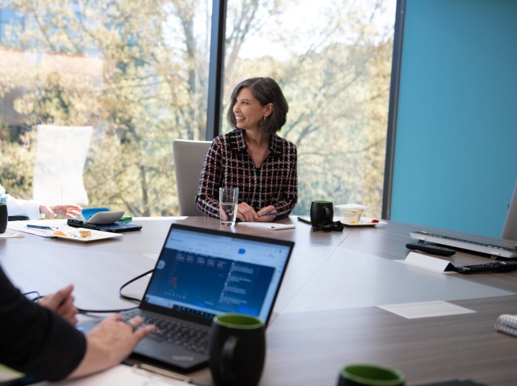 Jeanne sits at conference table, smiling while someone uses a computer in the foreground