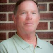 Man in green polo shirt standing in front of brick wall looking at camera