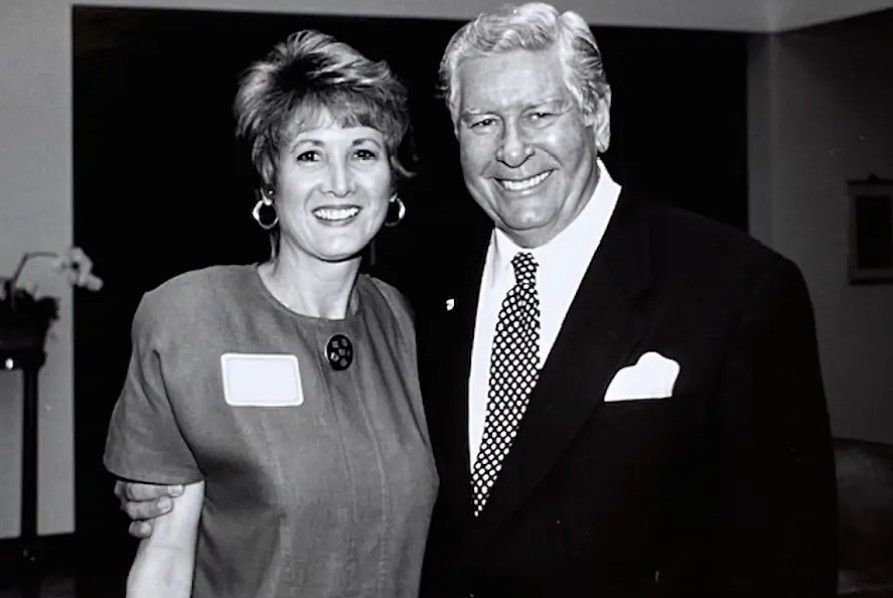 An archival black and white photo of a woman in a dress on the left posing for a picture with a gray-haired man in a suit.